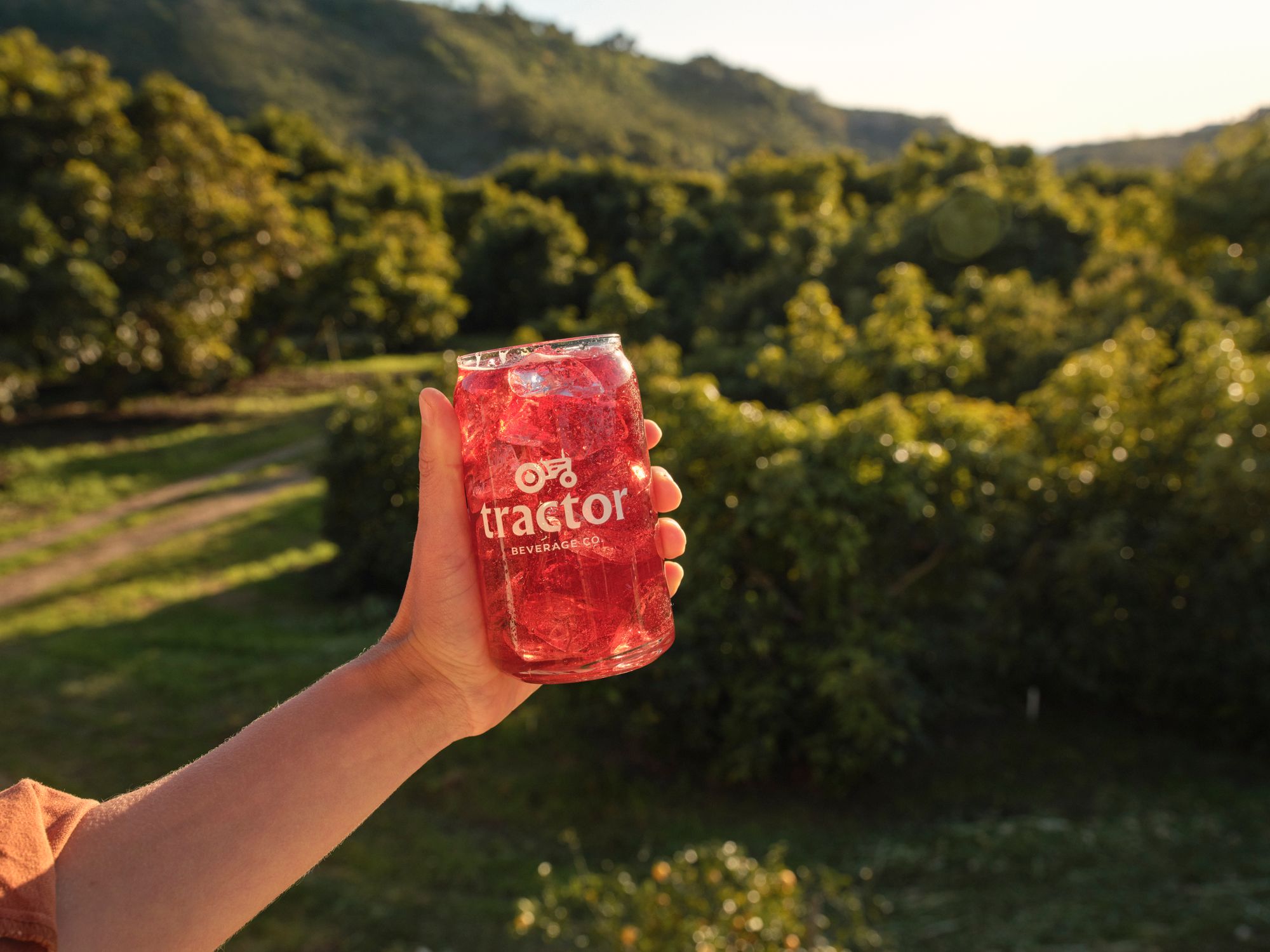 Person holdng a glass of a red beverage from Tractor Beverage co., with a scenic backdrop of trees and hills